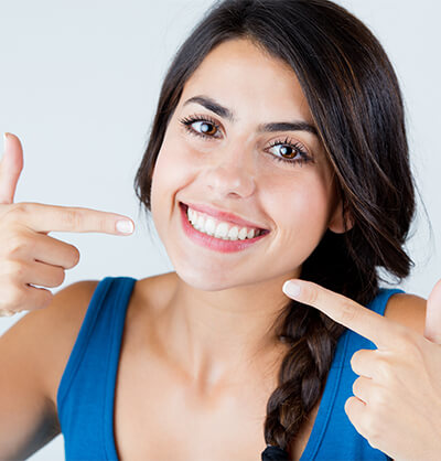 Brunette woman in blue tank top smiling and pointing to white teeth with both hands