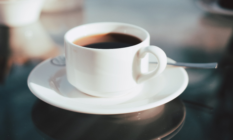 Closeup of a white mug and saucer with black coffee with caffeine next to a silver spoon